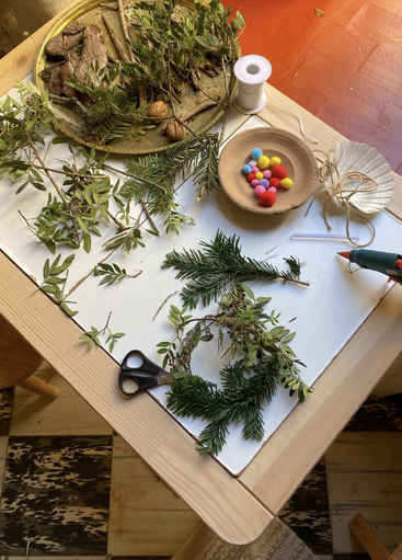 Une table en bois pour le bricolage, avec des ciseaux, un pistolet à colle, de la ficelle, des pompons colorés, des coquillages, de la verdure assortie, des brindilles et des noix disposés dans des bols et des plateaux.