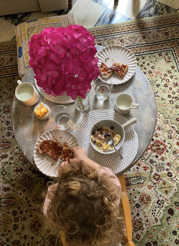 Un enfant prend son petit-déjeuner à une petite table ronde sur laquelle sont disposés des assiettes de pain grillé, des flocons d'avoine, des verres d'eau et un centre de table à fleurs rose vif sur un tapis à motifs.
