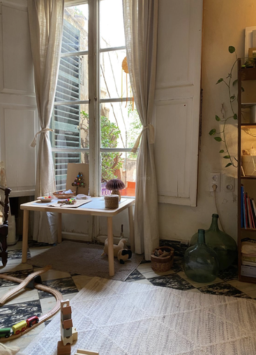 La lumière du soleil pénètre par les portes-fenêtres aux rideaux diaphanes. Une aire de jeu confortable et adaptée aux enfants comprend une petite table, des jouets, des rails de train en bois et un décor terreux et apaisant.