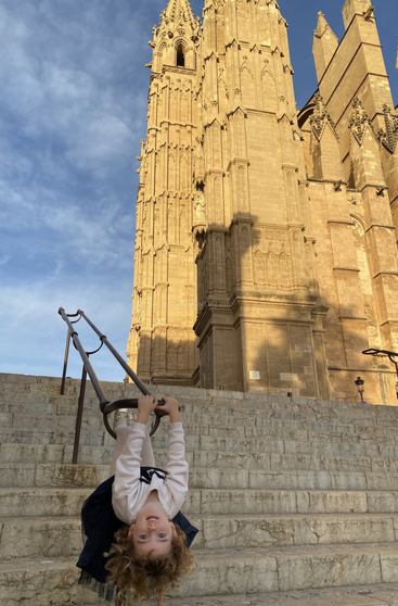 Un enfant est joyeusement suspendu à la rampe d'un escalier, le sourire aux lèvres. Derrière eux se dresse une majestueuse cathédrale gothique éclairée par le soleil, sous un ciel bleu partiellement nuageux.