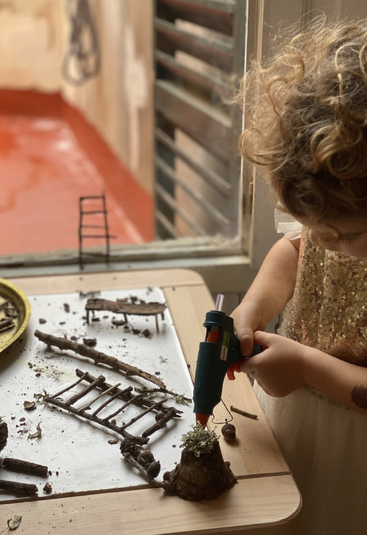 Un enfant vêtu d'une robe dorée utilise un pistolet à colle pour assembler des matériaux naturels tels que des bâtons et de la mousse, créant ainsi des structures miniatures sur une table près d'une fenêtre.