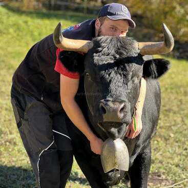 Ein Mann in Outdoor-Kleidung, der eine Mütze trägt, umarmt einen großen schwarzen Stier mit markanten Hörnern. Der Stier steht ruhig auf einer grasbewachsenen Wiese und trägt eine Glocke.