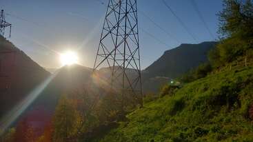 La photo immortalise un lever de soleil pittoresque derrière les montagnes, les rayons du soleil filtrant à travers un pylône électrique. Une herbe verte luxuriante et des arbres encadrent ce paysage vallonné paisible.