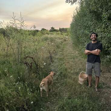 Un hombre sonriente está descalzo en un camino de hierba junto a un perro marrón y una cesta de mimbre, rodeado de exuberante vegetación bajo un cielo en tonos pastel al atardecer.