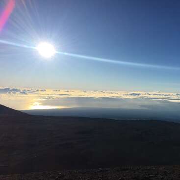 L'image représente un paysage serein avec une colline sombre, un soleil radieux et une vaste étendue de nuages, évoquant un sentiment de tranquillité et de beauté naturelle.