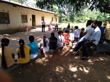Eine Gruppe von Menschen sitzt draußen im Schatten eines Baumes und hört aufmerksam zu, wenn jemand vor einem Gebäude spricht, was auf ein Gemeindetreffen oder eine Versammlung hindeutet.