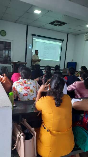 Ein Klassenzimmer mit einer Lehrkraft, die Informationen auf einer Projektionsfläche präsentiert, während die Schüler/innen aufmerksam zuhören und sich Notizen machen. Taschen und Wasserflaschen sind sichtbar.