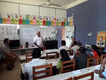 Das Bild zeigt ein Klassenzimmer mit einer Gruppe von Erwachsenen, die an Tischen sitzen und an einer Präsentation teilnehmen, die von einem Mann an der Stirnseite des Raumes gehalten wird.