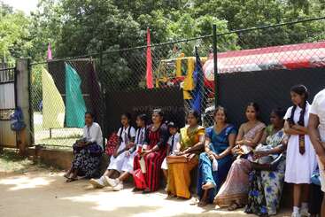 Das Bild zeigt eine Gruppe von Frauen und Kindern, die auf einer Bank sitzen, mit einem Maschendrahtzaun und Bäumen im Hintergrund und einem roten und gelben Fahrzeug hinter ihnen.