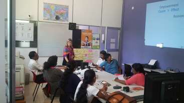 Das Bild zeigt eine Frau, die vorne in einem Klassenzimmer steht und eine Gruppe von Schülern anspricht, die um einen Tisch herum sitzen und lernen.