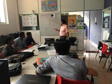 Um homem está em frente a um quadro branco, ensinando um grupo de alunos sentados em uma mesa, com laptops e papéis espalhados pela sala.