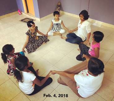 Das Bild zeigt eine Gruppe von Kindern und einen Erwachsenen, die in einem Kreis auf dem Boden sitzen und sich an den Händen halten, in einem Raum mit lila Wänden und beigen Fliesen.