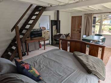Cette chambre rustique et chaleureuse dispose d'un escalier en bois, d'un lit, d'une petite télévision, d'un poêle à bois, d'une table à manger, d'un coin cuisine et de grandes fenêtres éclairées par la lumière naturelle.
