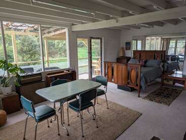 Cette image montre un espace de vie ouvert et confortable avec une table à manger vintage, des chaises vertes, un lit en bois, de grandes fenêtres, de la lumière naturelle et une abondante verdure à l'extérieur.