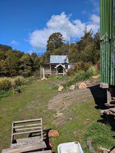 Une petite cabane rustique au toit de tôle se trouve dans une clairière verdoyante et boisée, sous un ciel bleu vif parsemé de nuages.