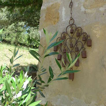 L'image représente un carillon éolien rustique en métal avec des cloches suspendues à un cadre en forme de cœur, accroché à un mur de pierre au milieu d'une végétation luxuriante.
