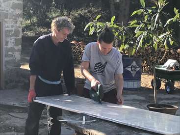 Deux personnes se trouvent à l'extérieur et travaillent sur une pièce de métal à l'aide d'une scie. Elles portent des vêtements décontractés et des gants, dans une cour avec des arbres et une brouette à proximité.