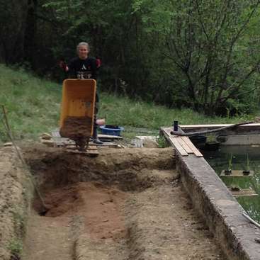 L'image représente un homme debout derrière une brouette remplie de terre, située près d'un étang et entourée d'arbres et d'herbe.