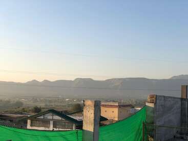 Cette image montre une vue panoramique sur des montagnes lointaines sous un ciel dégagé, avec au premier plan des toits, des filets verts, des piliers en béton et quelques maisons. L'atmosphère est paisible.