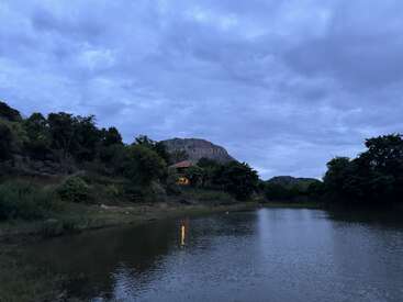 A serene landscape at dusk shows a calm lake reflecting the cloudy blue sky, surrounded by trees, hills, and a cozy house with warm lights.