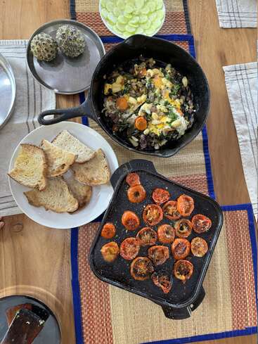 A rustic meal with toasted bread, roasted tomatoes, a hearty vegetable and bean skillet, two custard apples, and sliced cucumbers, beautifully arranged on a wooden table.