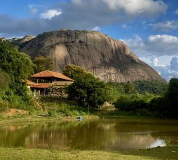 A serene landscape featuring a large rocky hill, a cozy house with a red roof, lush green trees, and a reflective pond under a partly cloudy sky.