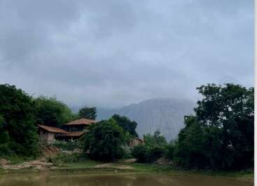 A rustic house with a red roof nestles among lush green trees by a calm river, misty mountains and cloudy sky create a tranquil, serene atmosphere.