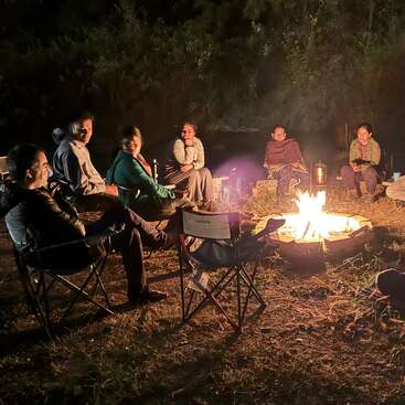 Um grupo de pessoas senta em círculo ao redor de uma fogueira quentinha à noite, curtindo a companhia uns dos outros ao ar livre, cercados pela escuridão e pela natureza.