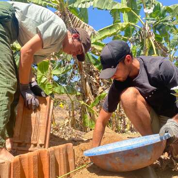 Duas pessoas estão trabalhando ao ar livre, cercadas por bananeiras. Elas estão usando luvas e chapéus, usando telhas e uma bacia azul, provavelmente construindo ou cuidando do jardim sob o sol.