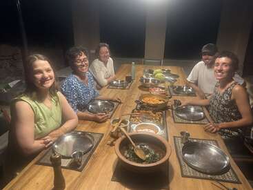 Cinco pessoas sentam-se em torno de uma mesa de madeira, sorridentes e prontas para o jantar. Comida deliciosa é servida em tigelas, criando uma atmosfera acolhedora, convidativa e comunitária.