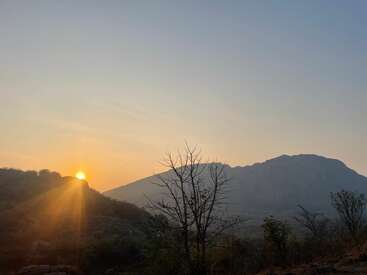 A imagem mostra um lindo nascer do sol sobre uma colina, com a luz quente do sol brilhando sobre uma paisagem tranquila de árvores e uma montanha distante sob um céu claro.