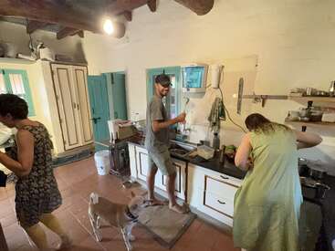 Três pessoas estão ocupadas preparando comida em uma cozinha rústica. Um cachorro fica por perto, observando-as. A sala tem portas azul-petróleo, vigas de madeira e prateleiras abertas.