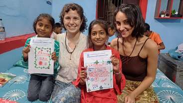 Zwei junge Mädchen zeigen stolz bunte Zeichnungen, während sie neben zwei lächelnden Frauen auf einem Bett in einem warm dekorierten Zimmer sitzen. Es ist ein fröhlicher, herzerwärmender Moment.