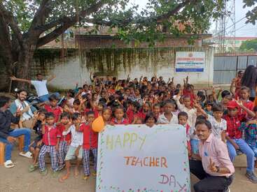 Eine Gruppe fröhlicher Kinder und ein paar Erwachsene feiern den Teacher's Day im Freien. Sie halten ein geschmücktes Schild unter einem großen Baum, lächeln und posieren zusammen.