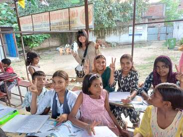 Eine Gruppe lächelnder Kinder sitzt an einem Tisch im Freien und lernt mit Heften. Sie machen Friedenszeichen, während eine fröhliche Frau hinter ihnen steht und zum Lernen auffordert.