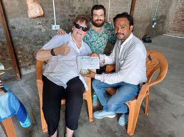 Drei Personen sitzen auf Plastikstühlen in einem rustikalen Raum. Eine Person hält eine Urkunde in der Hand, eine andere zeigt den Daumen hoch. Alle scheinen glücklich und stolz auf ihre Leistung zu sein.