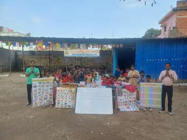 A group of children and adults gather outside a rural school for a learning event, surrounded by educational charts, a whiteboard, and colorful decorations.