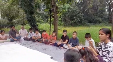 Um grupo de crianças e dois adultos sentados de pernas cruzadas em um tapete ao ar livre, cercado por árvores e vegetação, participando de uma aparente discussão ou atividade em grupo.