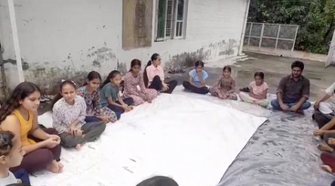 Um grupo de pessoas, a maioria meninas e alguns homens, está sentado em um círculo ao ar livre sobre grandes lençóis brancos perto da parede de um prédio.