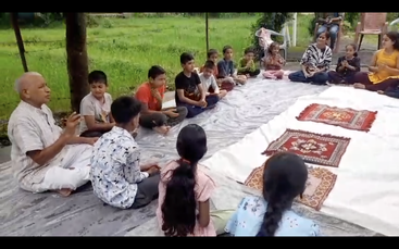 Um grupo de crianças senta-se com as pernas cruzadas em uma esteira ao ar livre, ouvindo atentamente um homem idoso, enquanto as mulheres supervisionam e a vegetação as rodeia.