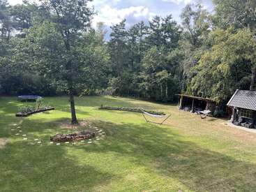 Une grande cour arrière avec une pelouse verte, un grand arbre, des parterres de fleurs, un hamac, un trampoline, un abri en bois, le tout entouré d'une forêt dense sous un ciel partiellement nuageux.