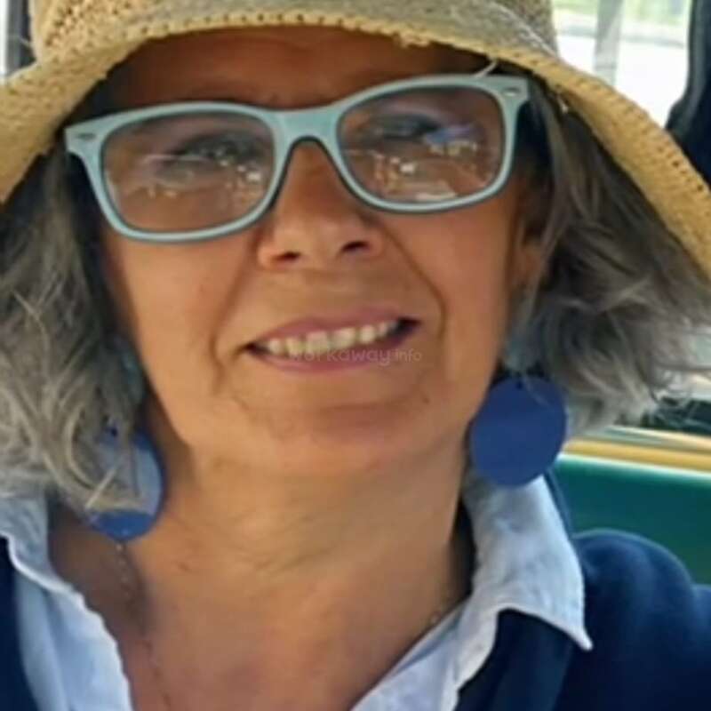 Eine Frau mit grauen Haaren hat einen breiten Strohhut, eine hellblaue Brille, große blaue Ohrringe und ein weißes Hemd unter einem blauen Pullover an und lächelt freundlich.