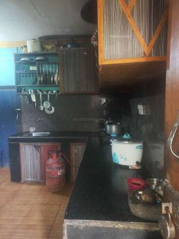 This is a modest kitchen with black countertops, cabinets, a gas cylinder, utensils rack, rice cooker, pots, and various kitchen items arranged neatly.