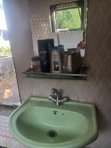 A green washbasin with a silver faucet is mounted below a mirror. On the glass shelf, toiletries like shampoo, conditioner, and a metal jug are placed.