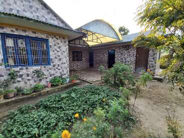 The image shows a house with stone-patterned walls, blue window grills, potted plants, a small courtyard, garden with greenery, flowers, and a covered entrance.