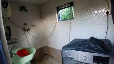 This image shows a small tiled bathroom with a green sink, red jug, washing machine, shower, window with greenery outside, and an electric water heater.
