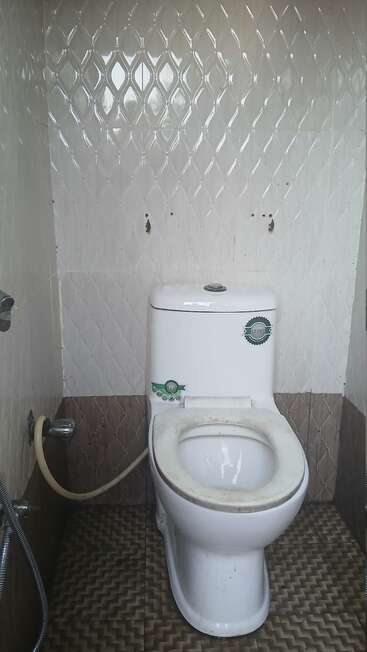 This image shows a white toilet with a dirty seat, tiled walls and floor, a handheld bidet sprayer, and a generally unclean appearance in a restroom.
