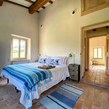 The image depicts a serene bedroom with a bed, nightstand, and rug, featuring white walls, wooden beams, and a window, exuding a sense of calm and tranquility.