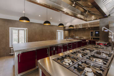 The image depicts a commercial kitchen with stainless steel appliances and countertops, featuring a large island and a sink, likely used for food preparation and cooking. The kitchen is well-organized and clean.