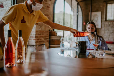 A person is pouring wine from a bottle into a glass, with a table and chairs in the background. The scene is set against a neutral-colored wall. The overall atmosphere is one of relaxation and leisure.